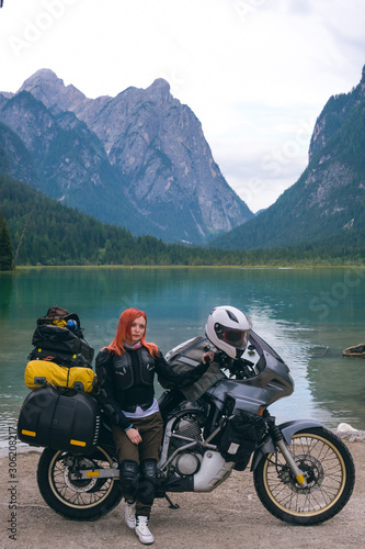 Vertical photo. Girl motobiker in protective turtle jacket stands with touring motorcycle. Extreme travel vacation, motorcyclist adventure lifestyle. Toblacher See, (Lago di Dobbiaco) Italy.