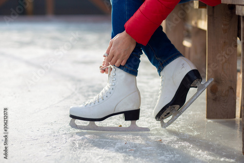 A girl in a red jacket is tying shoelaces on figured white skates on an ice rink photo