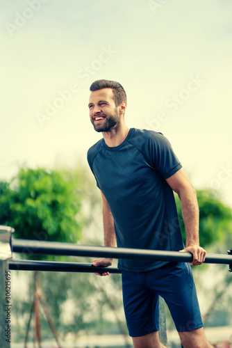 Happy smiling handsome bearded man, doing push ups exercise at horizontal bars, during workout training, outdoors. Fitness, sport, exercising, crossfit and workout concept.