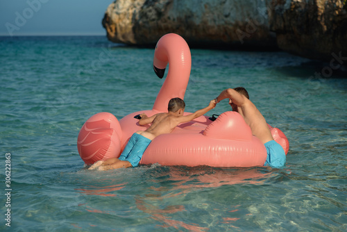 European man is happy to spend his summer holidays with his son. They are swimming in the ocean on inflatable pink flamingo and enjoying pastime.