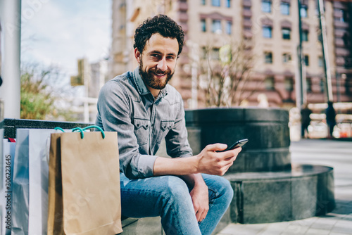 Φωτογραφία Portrait of cheerful caucasian bearded guy satisfied with delivery using app on mobile phone sitting with packages outdoors, smiling male buyer holding smartphone looking at camera on street