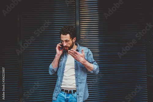 Angry bearded young man gesturing hands while solving problem during phone conversation on smartphone.Annoyed hipster blogger has quarrel while calling on mobile phone standing outdoors against wall
