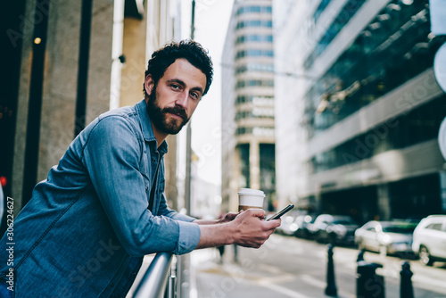 Portrait of bearded young man dressed in casual wear holding smartphone in hand and chatting online in social networks.Hipster guy in denim wear installing app on cellular standing on balcony