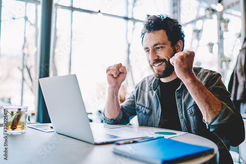 Overjoyed caucasian hipster guy celebrating victory in computer game enjoy playing on laptop computer during free time, happy male freelancer excited with completing online project successfully.