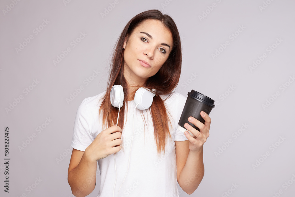 Serious female student keeps hand on wire, has tired expression, tries to refresh herself with strong coffee and loud music, wears mock up white t shirt, stands indoor with free space aside on grey. Serious female student keeps hand on wire, has tired expression, tries to refresh herself with strong coffee and loud music, wears mock up white t shirt, stands indoor with free space aside on grey.