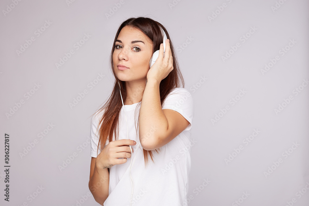 Young pensive pleasant looking redhead lady, holds earphone, makes song playlist, uses special app, wears white t-shirt, focuses at camera, being deep in thoughts, isolated on grey studio blank wall. Young pensive pleasant looking redhead lady, holds earphone, makes song playlist, uses special app, wears white t-shirt, focuses at camera, being deep in thoughts, isolated on grey studio blank wall.