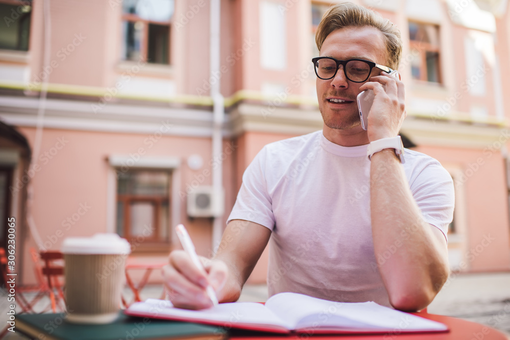 Clever male student in optical eyewear learning in public place preparing to university examination outdoors, successful man in classic glasses for vision correction having roaming cell call