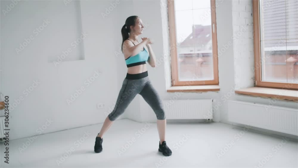 Young fit brunette girl with pigtail in blue top, gray leggings , black sneakers does exercises in bright room. Doing sports and fitness training at home. Healthy lifestyle. girl makes lunges, punches Young fit brunette girl with pigtail in blue top, gray leggings , black sneakers does exercises in bright room. Doing sports and fitness training at home. Healthy lifestyle. girl makes lunges, punches