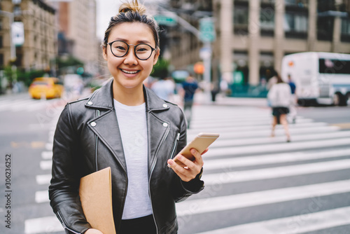 Half length portrait of cute emotional asian woman in eyewear spending leisure time walking at summer urban setting.Pretty smiling female person looking at camera while downloading app on phone
