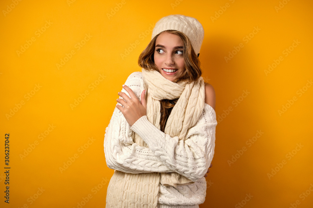 Beautiful girl with clean skin and loose curls in a white sweater and hat on a yellow background, wrapping herself in a scarf Beautiful girl with clean skin and loose curls in a white sweater and hat on a yellow background, wrapping herself in a scarf