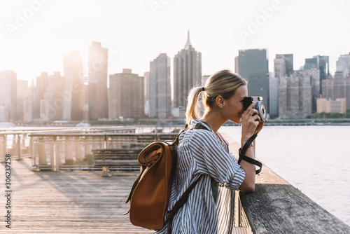 Side view of skilled hipster girl with instant camera clicking picture of city landscape standing at urban setting with Manhattan view, positive female tourist with backpack enjoying getaway