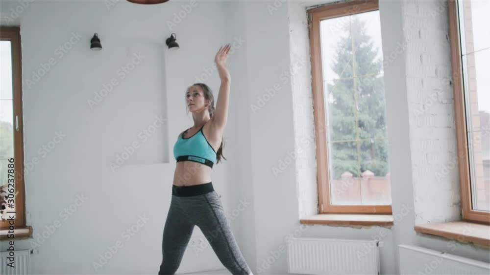 Young fit sporty pretty brunette girl with pigtail in blue top, gray leggings does sport exercises in bright room. Doing fitness training at home. Healthy lifestyle, healthcare. stretching, leaning Young fit sporty pretty brunette girl with pigtail in blue top, gray leggings does sport exercises in bright room. Doing fitness training at home. Healthy lifestyle, healthcare. stretching, leaning