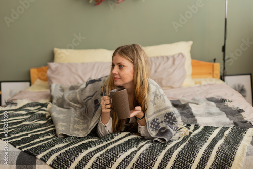 Beautiful woman on the bed under a plaid drinks tea. Modern and stylish room design.