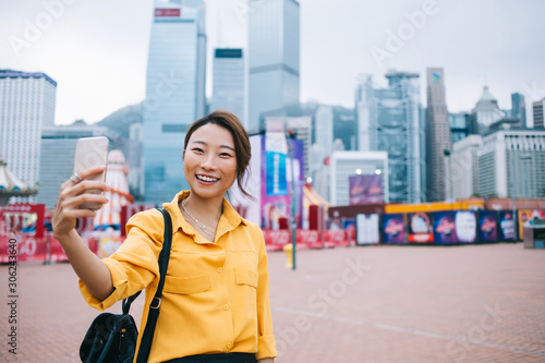 Half length portrait of cheerful female influencer clicking selfie pictures for sharing to social networks during Chinese holidays for exploring Hong Kong, happy hipster girl photographing herself