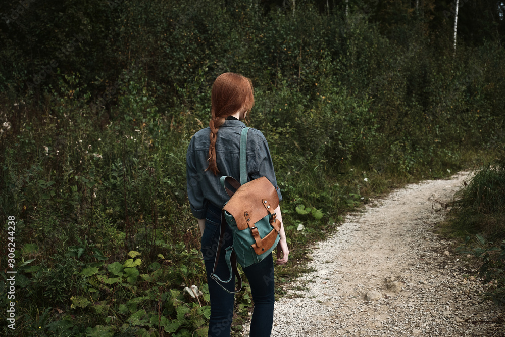 A red-haired girl is walking in the forest along the road with a backpack, a frame from the back from the side. A red-haired girl is walking in the forest along the road with a backpack, a frame from the back from the side.