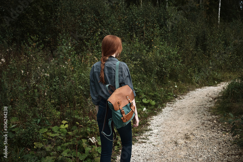 A red-haired girl is walking in the forest along the road with a backpack, a frame from the back from the side.