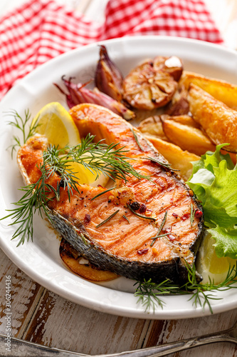 Grilled salmon steak, a portion of grilled salmon with fresh lettuce and potato wedges on a white ceramic plate on a wooden rustic table, close-up. Grilled seafood.