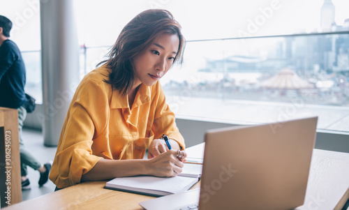 Asian female writing and looking at screen display at workplace photo