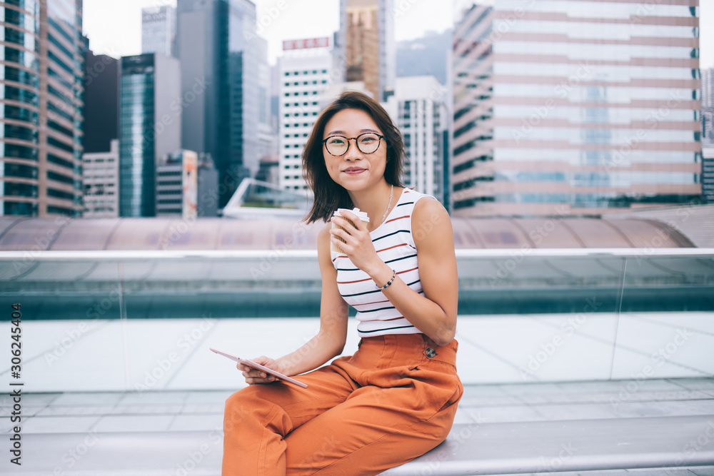 Fototapeta premium Portrait of Chinese female blogger with digital tablet for networking in hand looking at camera and smiling during free time at urban setting, positive woman in optical glasses for vision correction