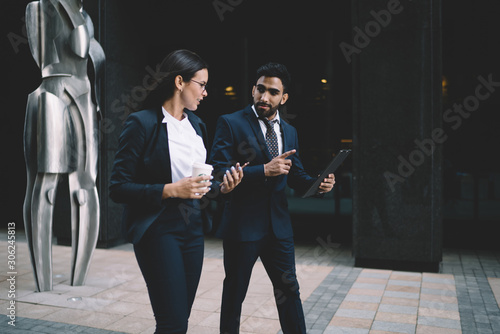 Two diverse mature economists dressed in formal suit walking around financial district with modern sculpture on background and discussing accounting report from folder on way to office building