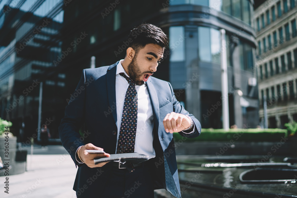 Spanish mature trader with folder in hand feeling shocked while checking  time on wearable wristwatch outdoors, amazed man dressed in formal wear  late for business meeting with financial expert foto de Stock