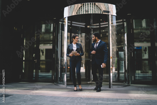Positive male and female coworkers going out from office building for spending break together enjoying friendship togetherness, successful economists in stylish suits discussing cooperation