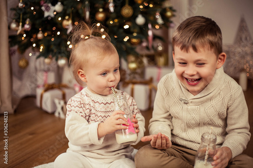 Little cute happy boy and girl sit near the Christmas tree and drink milk with Christmas cookies. Christmas. New Year.