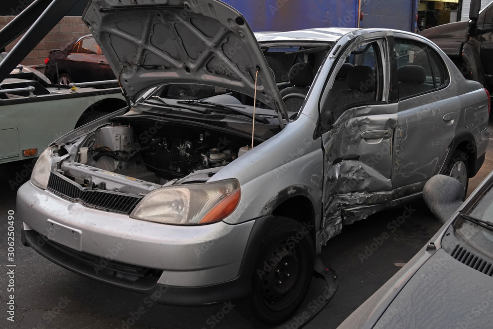 A generic passenger car with major damage to the driver side is shown in an automotive salvage yard during the late afternoon. A generic passenger car with major damage to the driver side is shown in an automotive salvage yard during the late afternoon.
