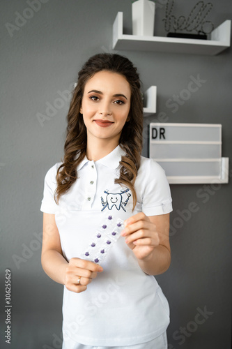 Young lady wearing on medical suit and posing at camera. Portrait of smiling dentist with brown hair. Happy doctor holding tools in hands. vertical photo