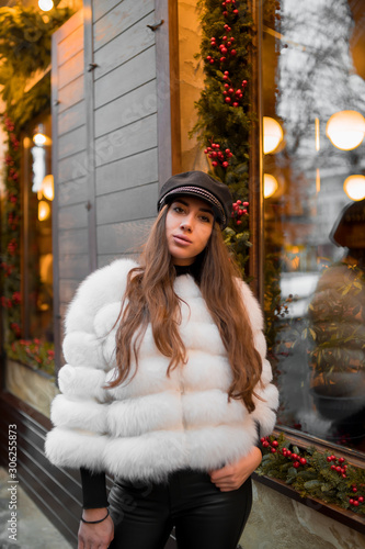 Beautiful young caucasian woman wearing natural short white fur coat posing for a portrait near Christmas showcase. Christmas decor. Fashion. Style. Winter clothes made of natural fur. Vertical