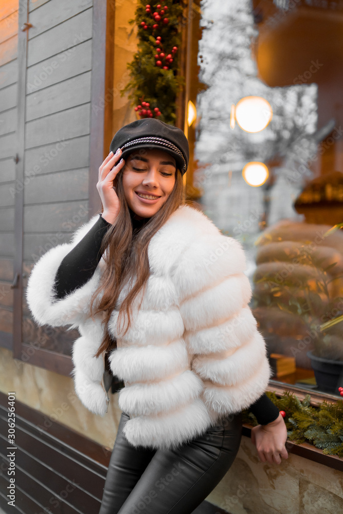 Beautiful young caucasian woman wearing natural short white fur coat posing for a portrait near Christmas showcase. Christmas decor. Fashion. Style. Winter clothes made of natural fur. Vertical