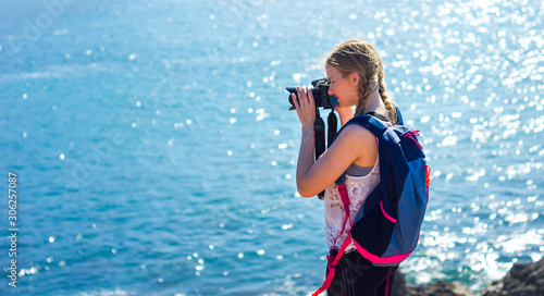 Young blonde caucasian girl taking photos of the ocean with backpack during vacation trip