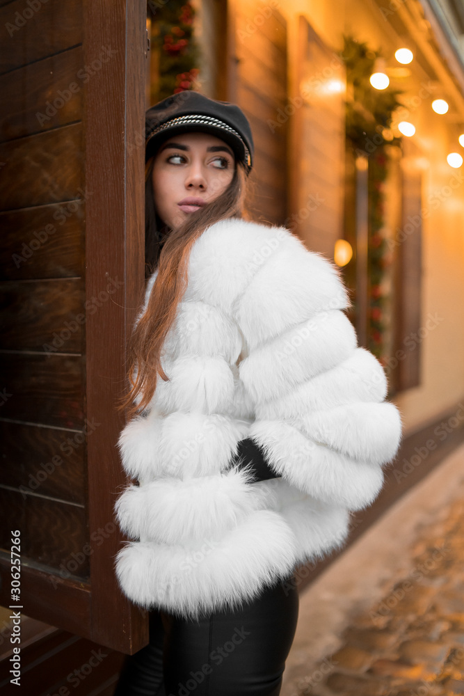 Beautiful young caucasian woman wearing natural short white fur coat posing for a portrait near Christmas showcase. Christmas decor. Fashion. Style. Winter clothes made of natural fur. Vertical
