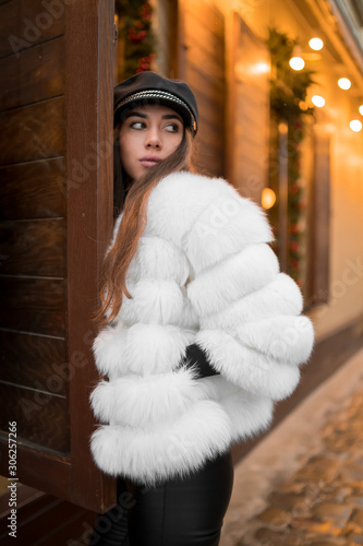 Obraz na płótnie Beautiful young caucasian woman wearing natural short white fur coat posing for a portrait near Christmas showcase. Christmas decor. Fashion. Style. Winter clothes made of natural fur. Vertical w salonie