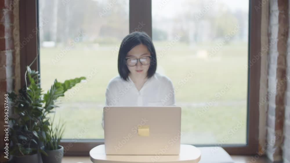 Zoom in of young attractive Asian businesswoman wearing white shirt and eyeglasses sitting on windowsill in café and working on laptop Zoom in of young attractive Asian businesswoman wearing white shirt and eyeglasses sitting on windowsill in café and working on laptop