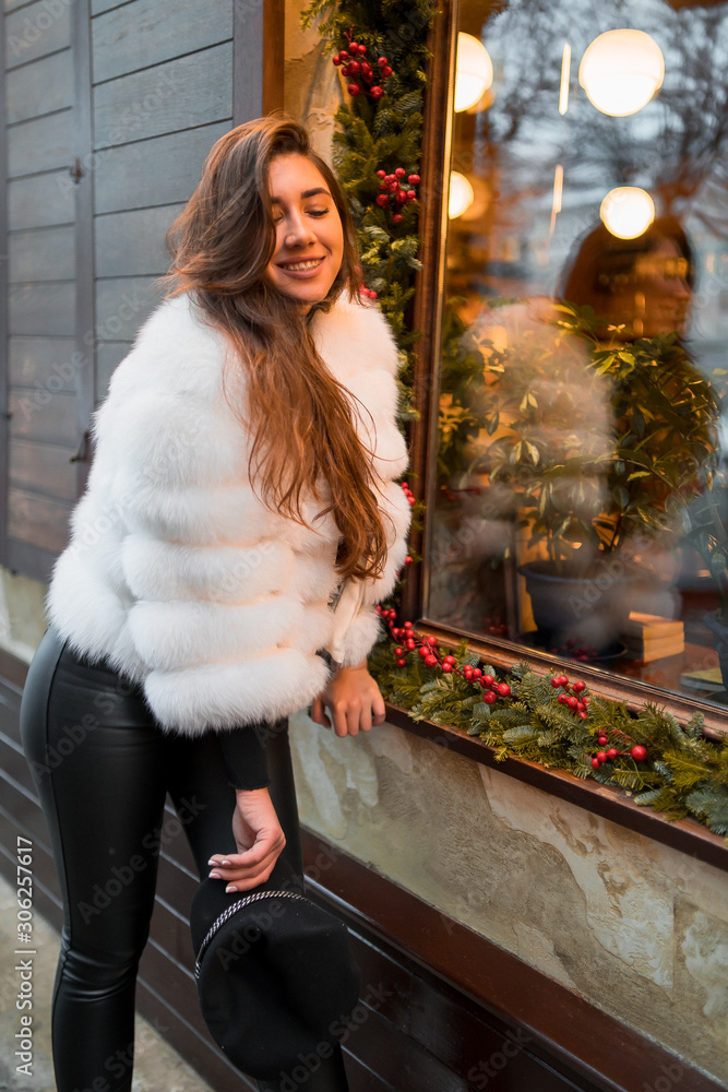 Beautiful young caucasian woman wearing natural short white fur coat posing for a portrait near Christmas showcase. Christmas decor. Fashion. Style. Winter clothes made of natural fur. Vertical