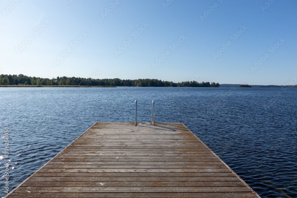 Obraz premium Wooden swimming dock pier with metal ladder on calm blue lake sunny day on nature finland idyllic natural resort