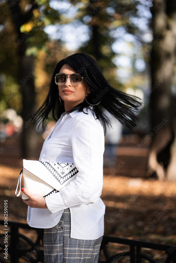 Obraz premium Autumn fashion outdoor. The girl in fashionable stylish white shirt , pants and sunglasses, autumnal lifestyle on the background of blurry yellow-green trees in the park. Vertical