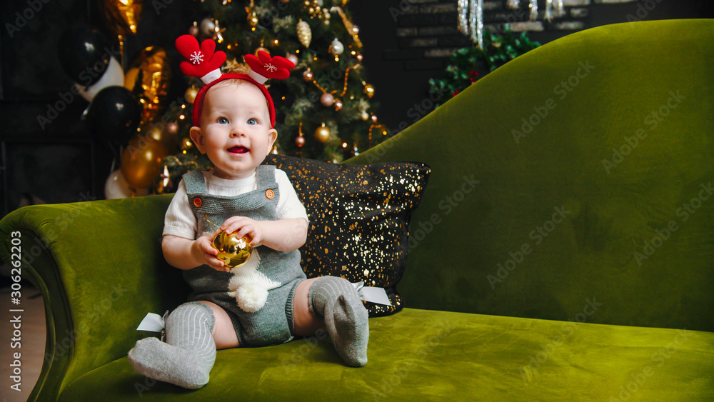 Christmas concept - smiling baby is sitting on the green sofa and playing with christmas ball