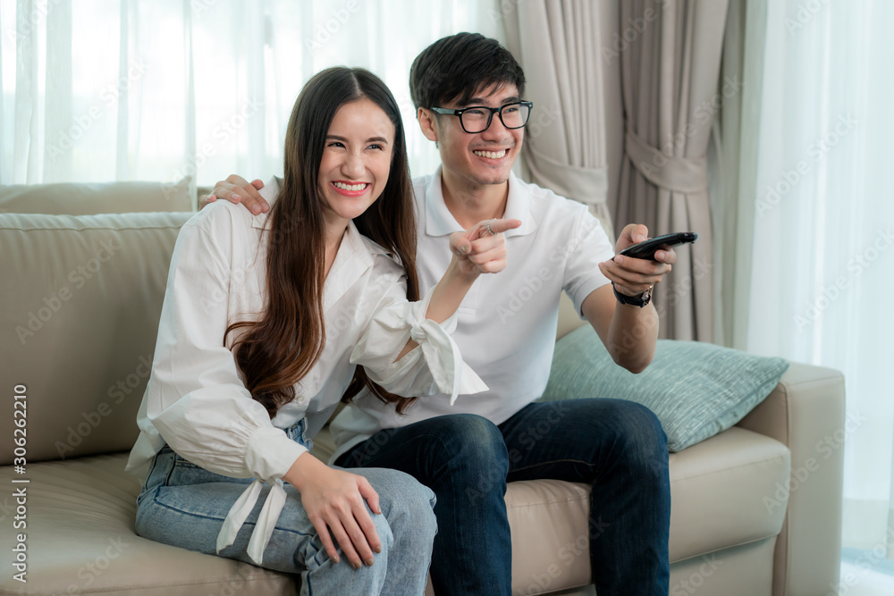 Asian young loving couple sitting and watching TV and smile when watching fun program in TV in a comfortable couch in the livingroom at home. Family lifestyle relax and recreation concept Asian young loving couple sitting and watching TV and smile when watching fun program in TV in a comfortable couch in the livingroom at home. Family lifestyle relax and recreation concept