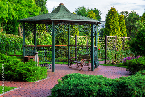 Vászonkép an iron gazebo with shingles and a park bench with bushes and trees, a lantern and an urn by the canopy on a summer day