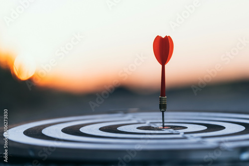 Close up red dart arrow hitting target center dartboard on sunset background. Business targeting and focus concept photo