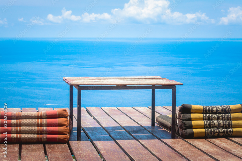 Fototapeta premium Table and Thai Triangle Pillows on Wooden Terrace with Tropical Beach, Blue Sky and Clouds in Background