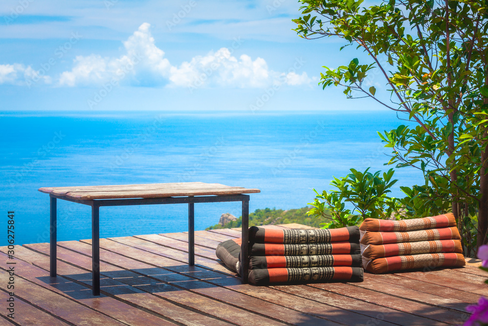 Table and Thai Triangle Pillows on Wooden Terrace with Tropical Beach, Blue Sky and Clouds in Background