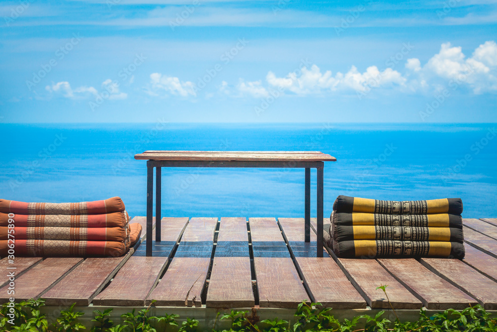 Table and Thai Triangle Pillows on Wooden Terrace with Tropical Beach, Blue Sky and Clouds in Background Table and Thai Triangle Pillows on Wooden Terrace with Tropical Beach, Blue Sky and Clouds in Background