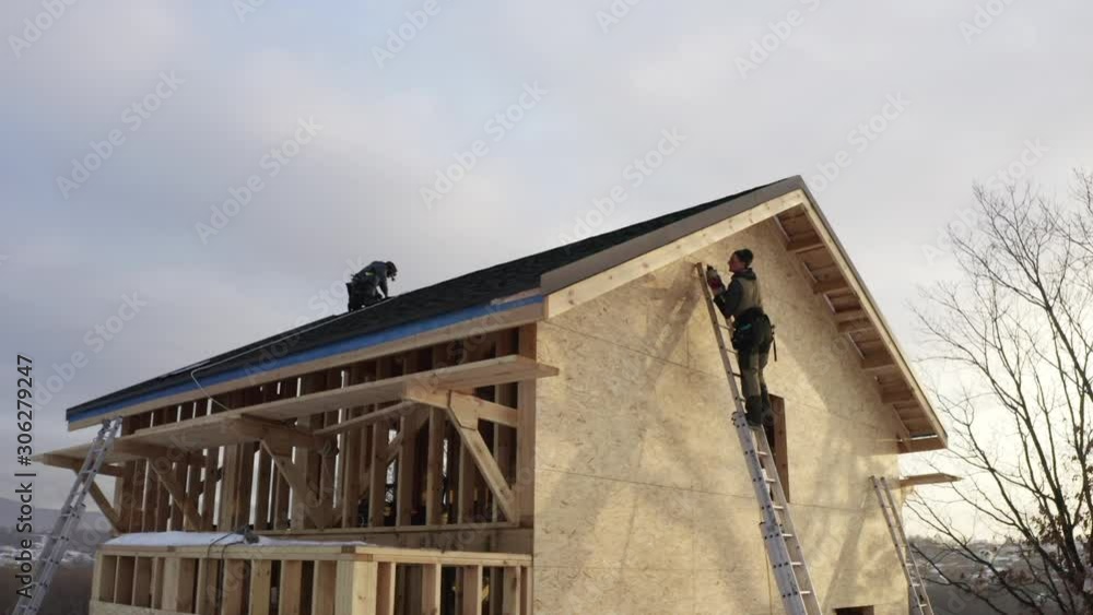 Aerial view of house under construction. One worker is covering the wooden frame with plywood using hydraulic hammer, another one is on the roof in safety harness Aerial view of house under construction. One worker is covering the wooden frame with plywood using hydraulic hammer, another one is on the roof in safety harness