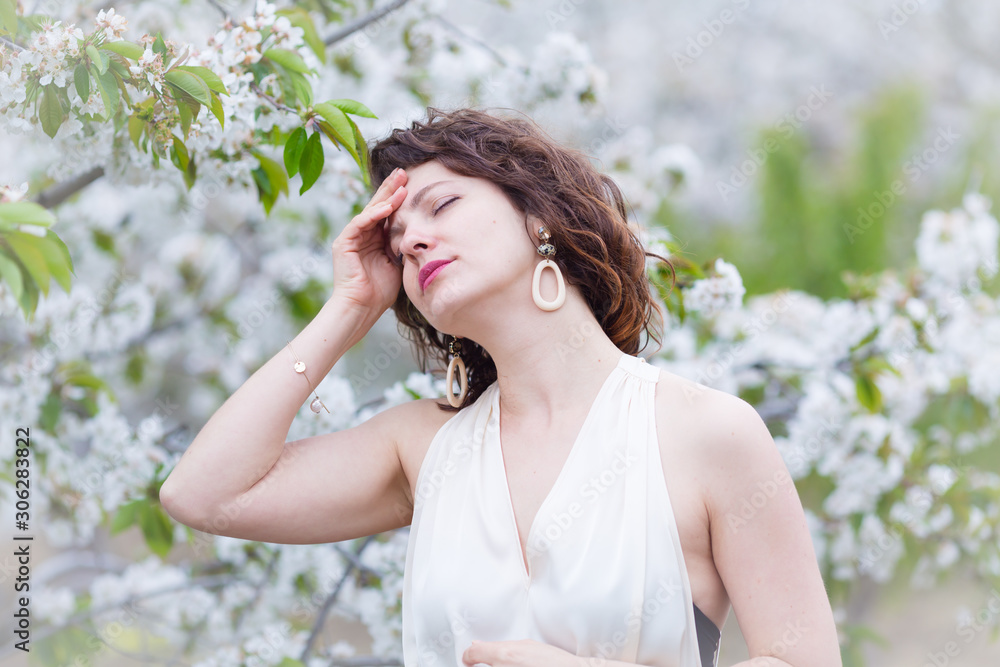 A portrait of Caucasian brunette woman near blossoming cherry tree. Painful emotions, headache