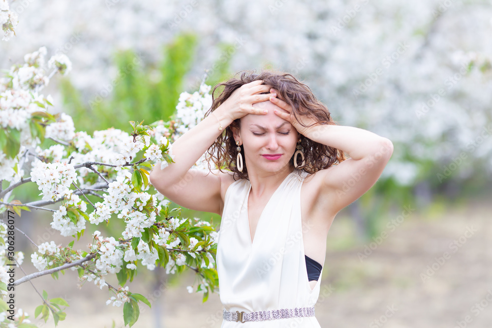 A portrait of Caucasian brunette woman near blossoming cherry tree. Painful emotions, headache