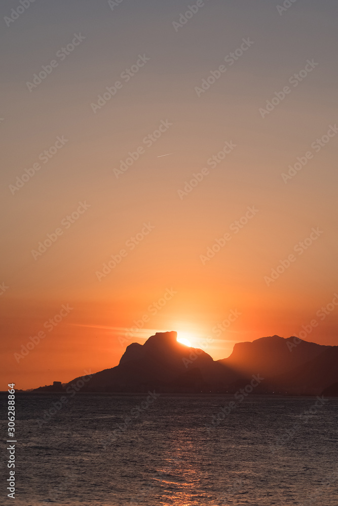 Orange sunset by the ocean in Piratininga, Niteroi, with sun dipping behing the Gavea Stone in Rio de Janeiro. A beach full of people can be seen in the foreground in shadow..