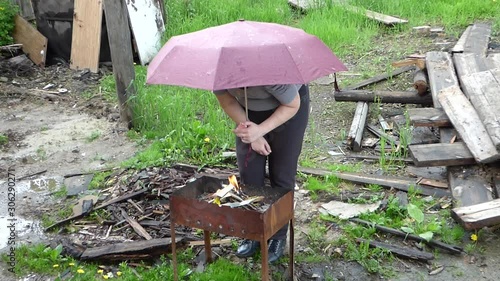 Women with umbrella light fire in barbecue under rain. Old dirty boards and grass around. Rural scene.